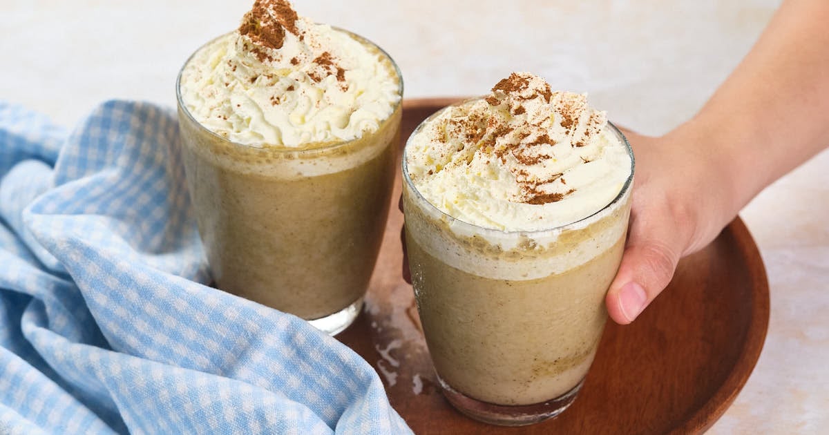 Two glasses of blended iced coffee topped with whipped cream and cinnamon sit on a wooden tray next to a blue checkered cloth; a hand holds one glass.