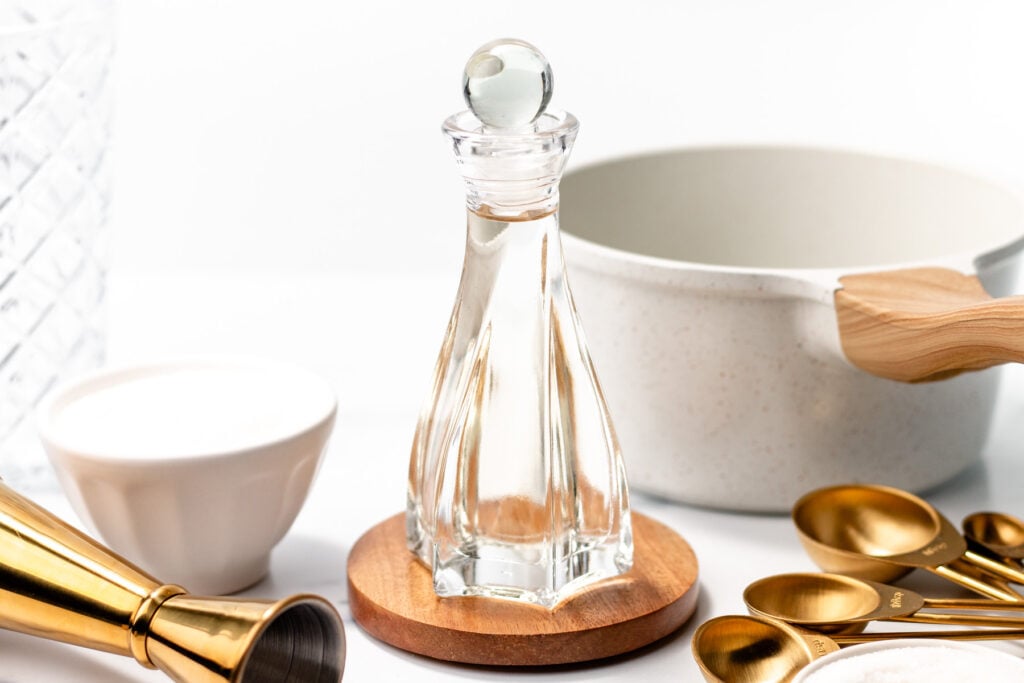 A glass cruet filled with clear liquid sits on a wooden coaster surrounded by measuring spoons, a white bowl, and a gold jigger on a white surface.