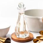 A glass cruet filled with clear liquid sits on a wooden coaster surrounded by measuring spoons, a white bowl, and a gold jigger on a white surface.