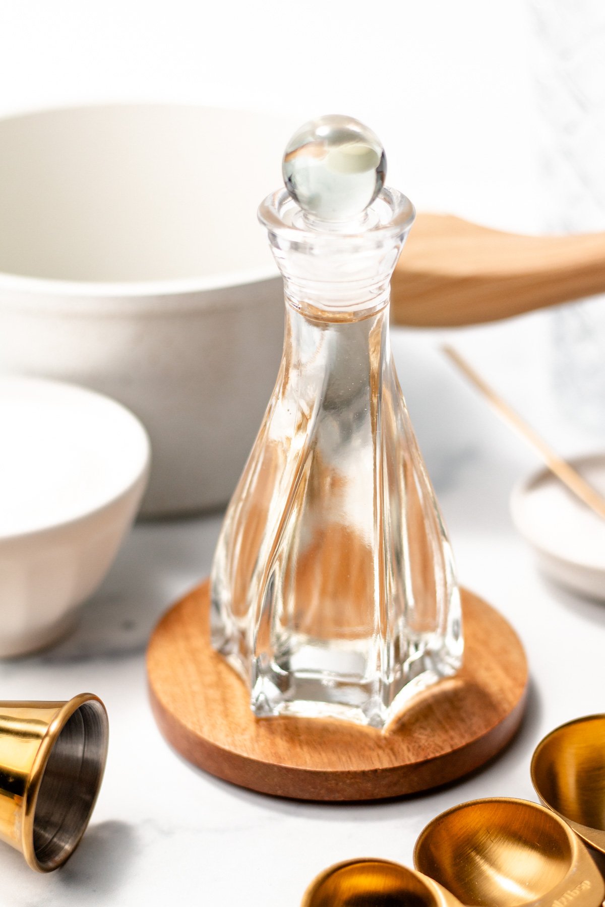 A clear glass decanter filled with liquid sits on a wooden coaster, surrounded by white bowls and gold measuring spoons on a white surface.