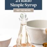 A glass bottle with clear liquid sits on a wooden coaster, surrounded by measuring cups and a white bowl; text reads “2:1 Ratio Rich Simple Syrup,” perfect as a cocktail sweetener.