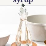 A glass bottle filled with clear Rich Simple Syrup sits on a wooden coaster, surrounded by a white bowl, mug, and gold measuring cups—perfect for elevating your favorite cocktail sweetener recipes.