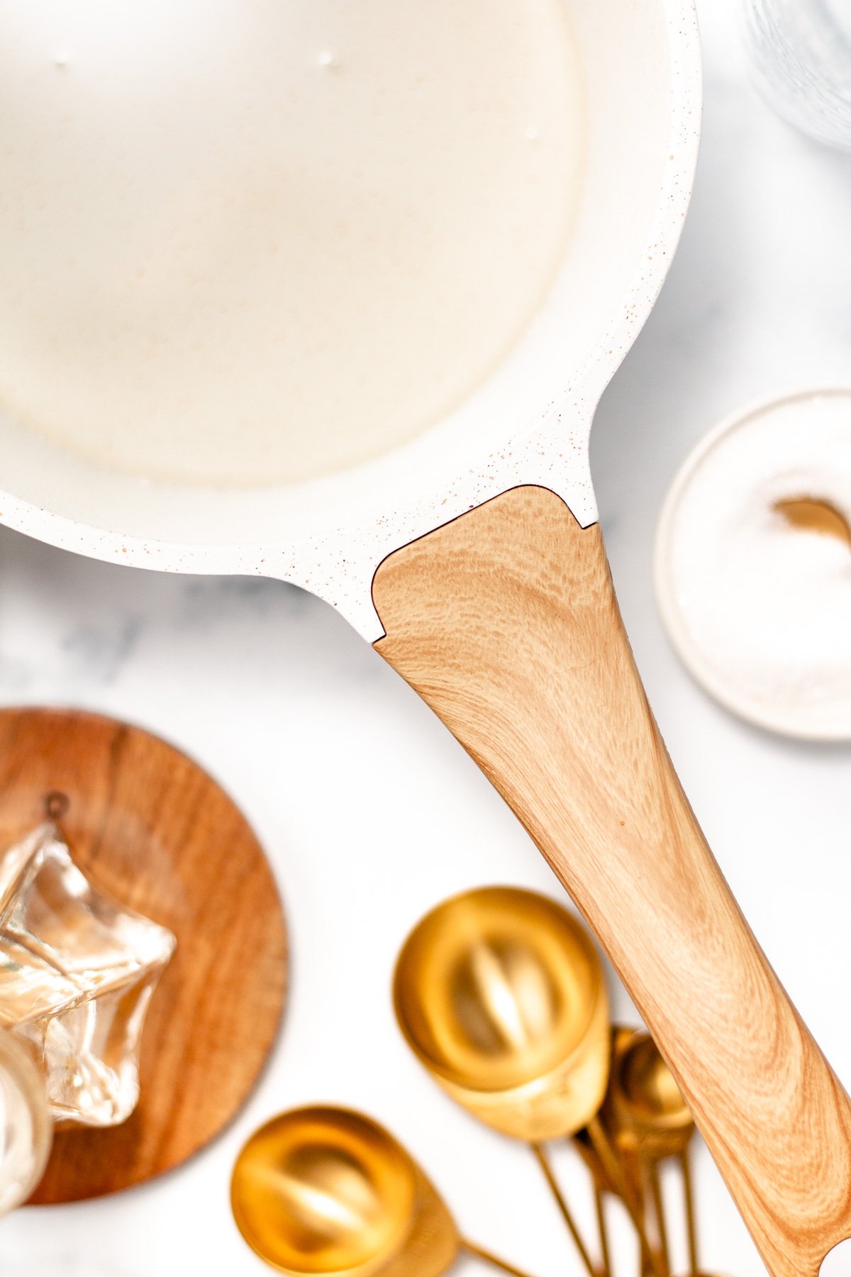White nonstick pan with a light wooden handle sits on a marble surface next to gold measuring spoons, a glass container, and a small bowl of salt.