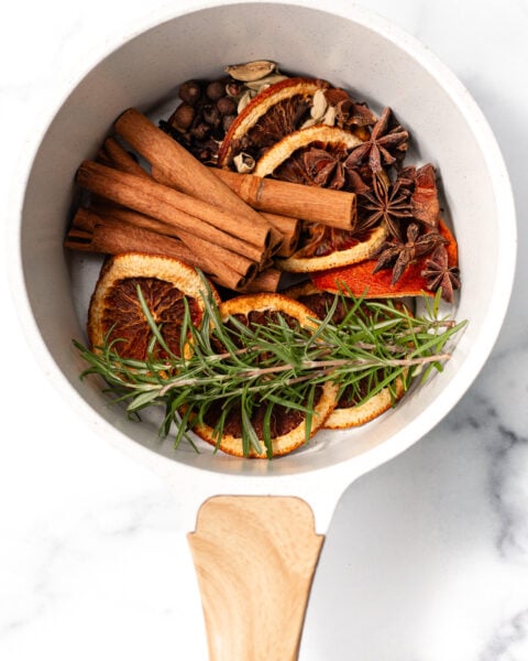 A white saucepan with dried orange slices, cinnamon sticks, star anise, peppercorns, cardamom pods, and sprigs of rosemary on a white marble surface.