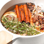 A white pot with cinnamon sticks, dried citrus slices, star anise, rosemary, and other spices as water is being poured in.