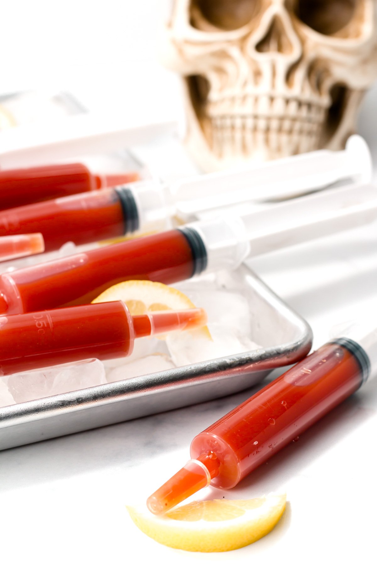 Red liquid-filled syringes rest on a tray of ice with lemon slices, with a skull decoration in the background.