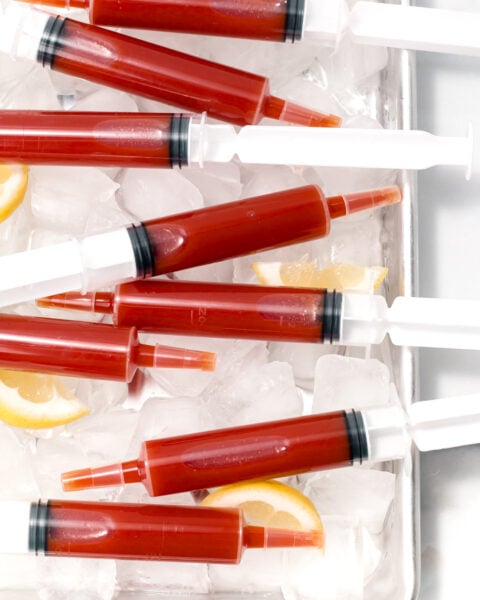 Red liquid-filled plastic syringes arranged on ice cubes with lemon slices on a metal tray.