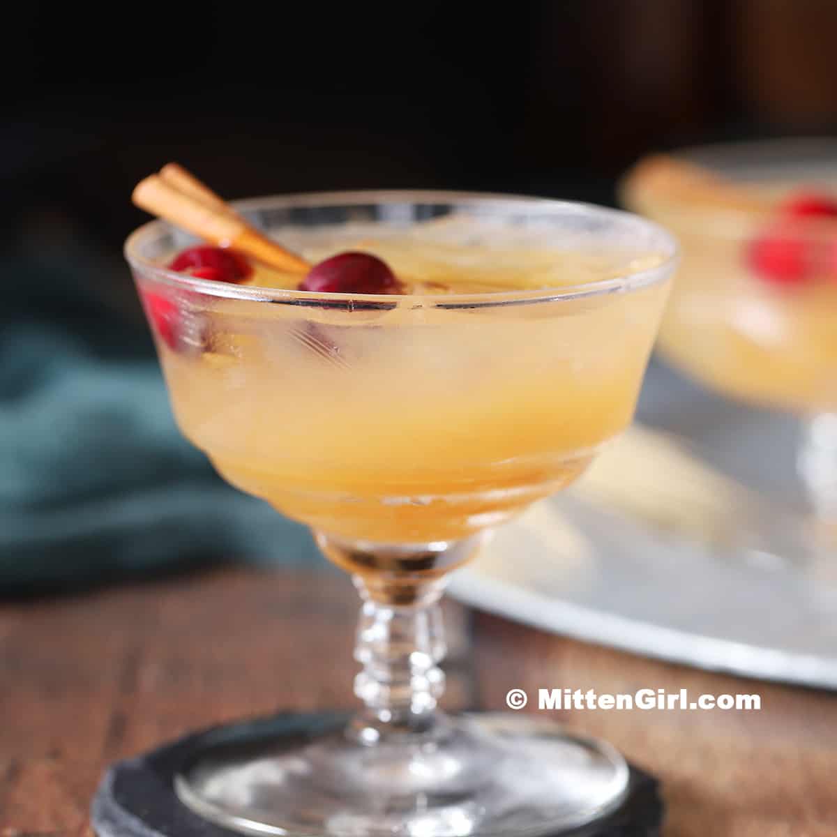 A coupe glass filled with a light orange cocktail, garnished with cherries and a cinnamon stick, sits on a dark coaster.