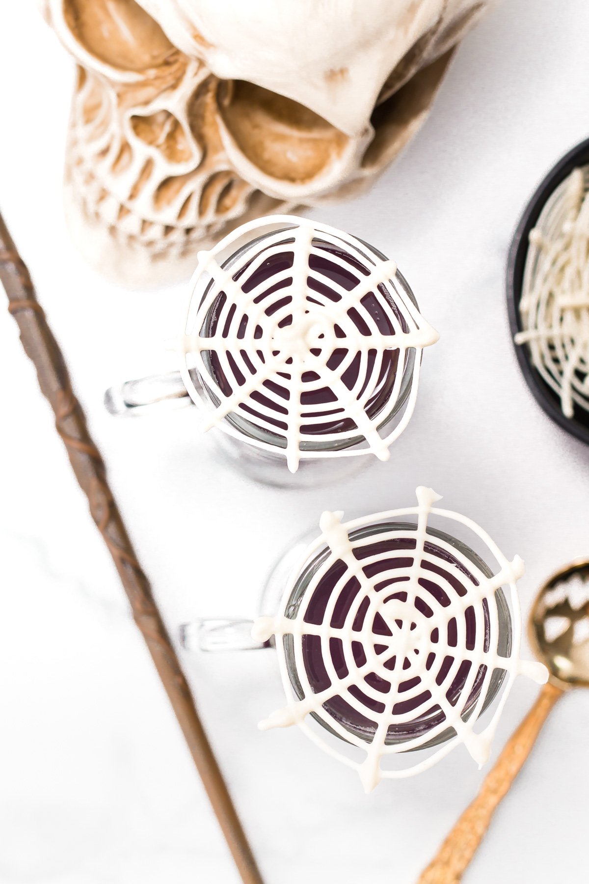 Two glass mugs with spiderweb designs on top, placed next to a skull decoration, a wand, and themed props on a white surface.