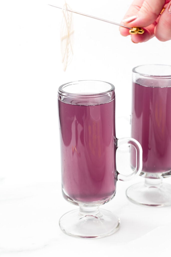 Two clear glass mugs filled with a purple liquid are shown. A hand holds a garnishing tool over one of the mugs. The background is white.