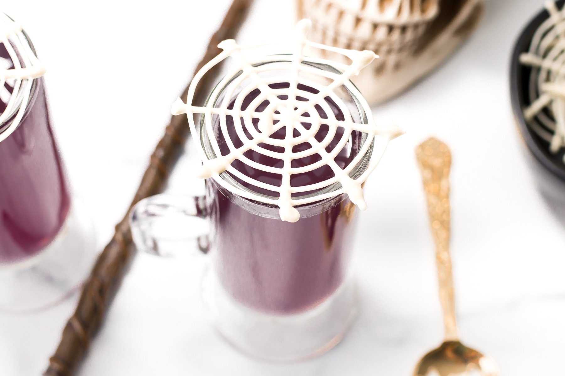 A glass mug filled with a dark purple drink, topped with a white chocolate spiderweb decoration, sits on a white surface next to a gold spoon and Halloween-themed props.
