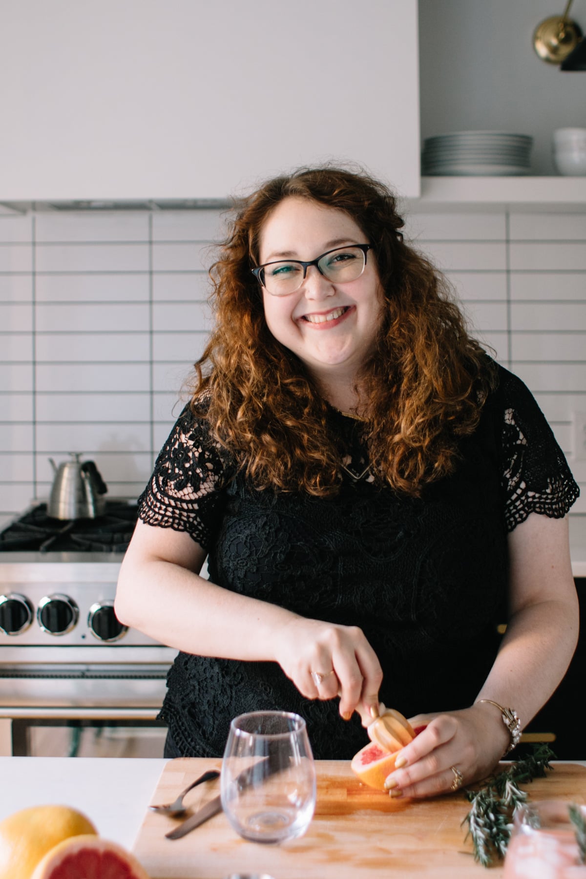 A woman with curly hair and glasses smiles while slicing a grapefruit in a modern kitchen, ready to craft refreshing holiday drinks from her new ebook.