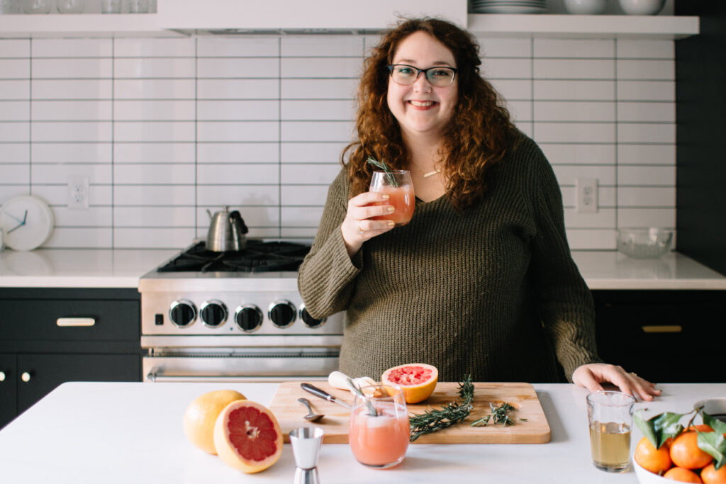A woman stands in a kitchen holding a glass of pink juice, with grapefruit, herbs, and kitchen tools on the counter in front of her&mdash;creating festive holiday drinks perfect for your next Holiday Spirits ebook.