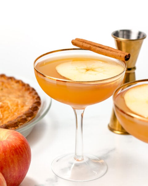 A cocktail garnished with an apple slice and cinnamon stick in a coupe glass, with apples, a pie, and a jigger in the background.