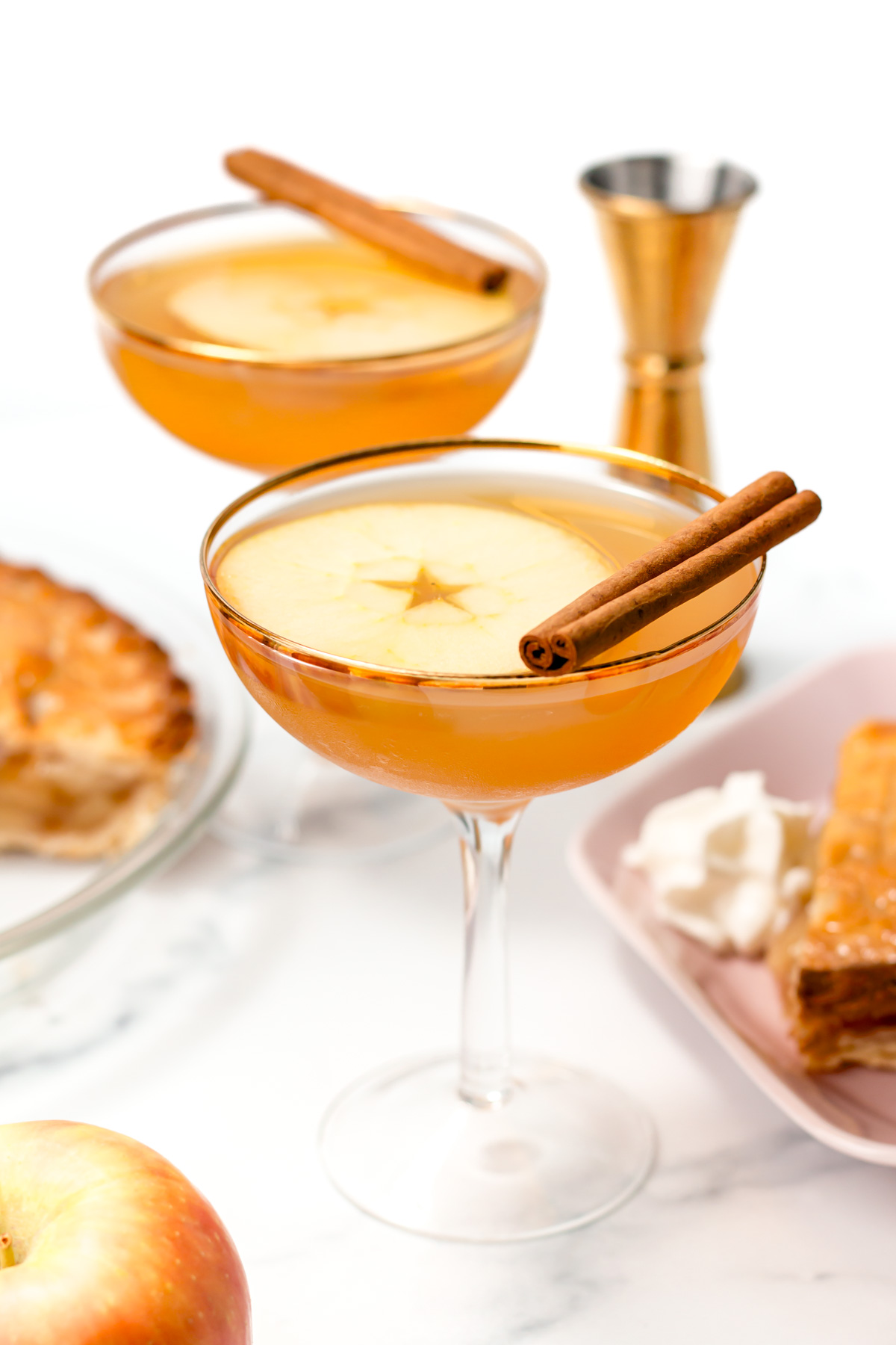 Two coupe glasses filled with an amber-colored drink, garnished with apple slices and cinnamon sticks, are set on a table with pie, an apple, and a jigger nearby.