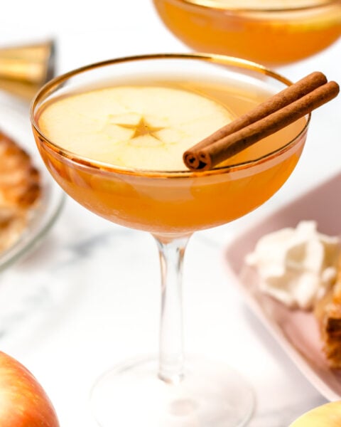 A cocktail in a coupe glass garnished with a thin apple slice and two cinnamon sticks, with additional apples and dessert in the background.