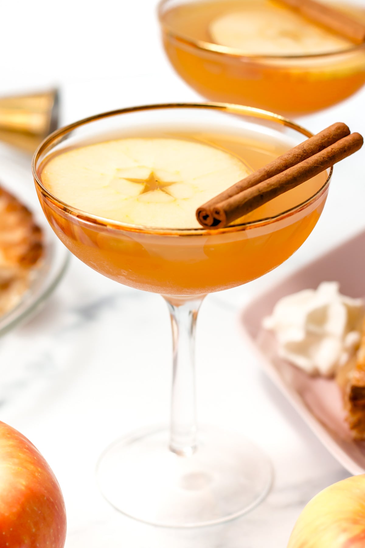 A cocktail in a coupe glass garnished with a thin apple slice and two cinnamon sticks, with additional apples and dessert in the background.