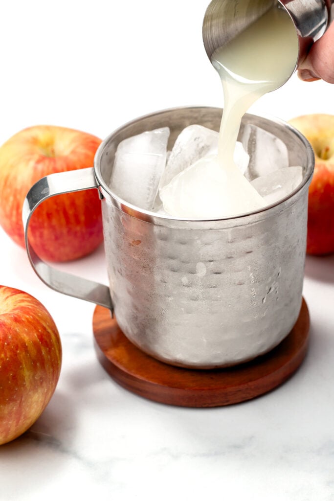 A metal cup filled with ice is being poured with a pale liquid, surrounded by apples on a white surface.
