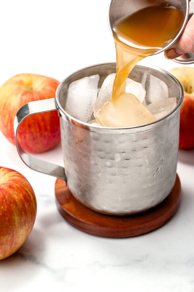A hand pours a brown liquid over ice cubes in a metal mug, with apples placed nearby on a white surface.