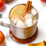 A metal mug filled with an iced beverage, garnished with a cinnamon stick and an apple slice, sits on a coaster surrounded by apples and a yellow cloth.