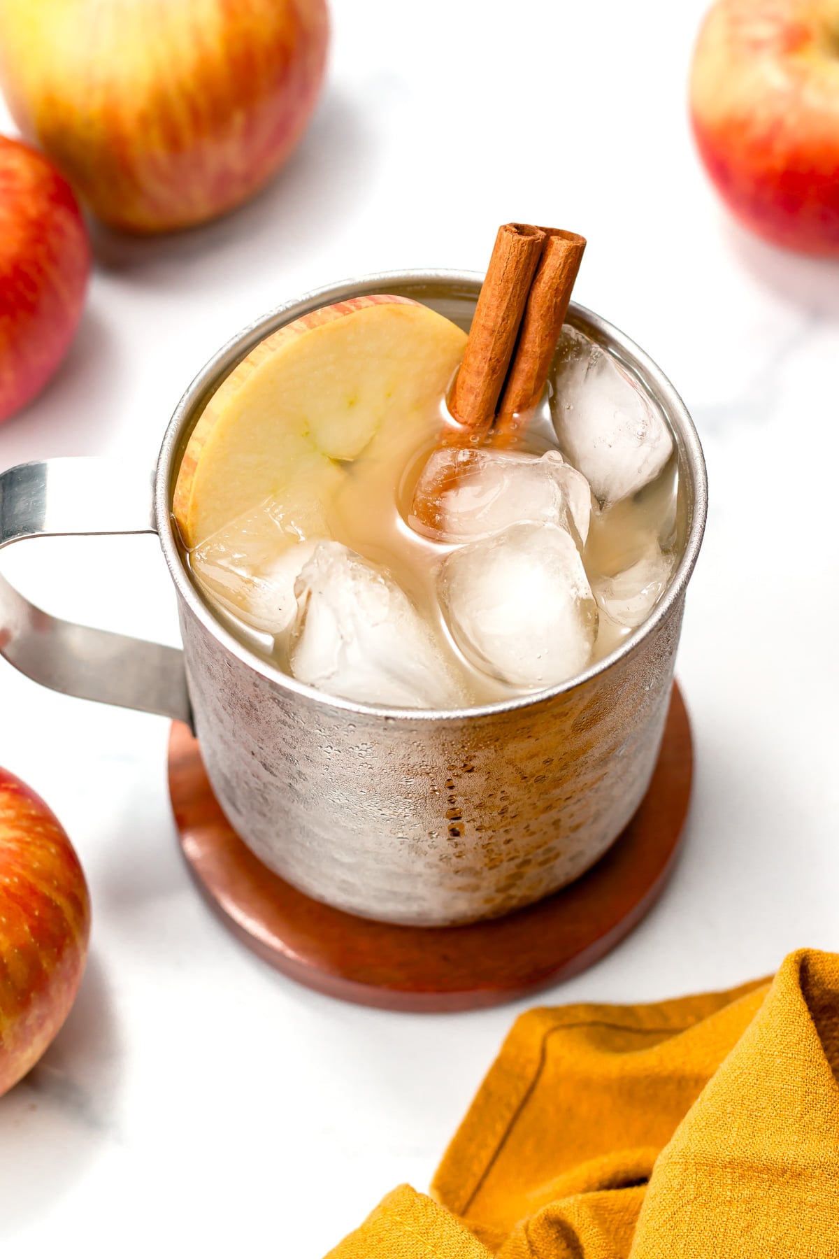 A metal mug filled with an iced beverage, garnished with a cinnamon stick and an apple slice, sits on a coaster surrounded by apples and a yellow cloth.