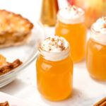 Three small mason jars filled with orange liquid, topped with whipped cream and cinnamon, sit on a white surface with apples, cinnamon sticks, and pie nearby.
