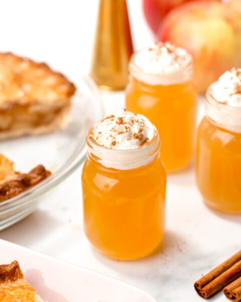 Three small mason jars filled with orange liquid, topped with whipped cream and cinnamon, sit on a white surface with apples, cinnamon sticks, and pie nearby.