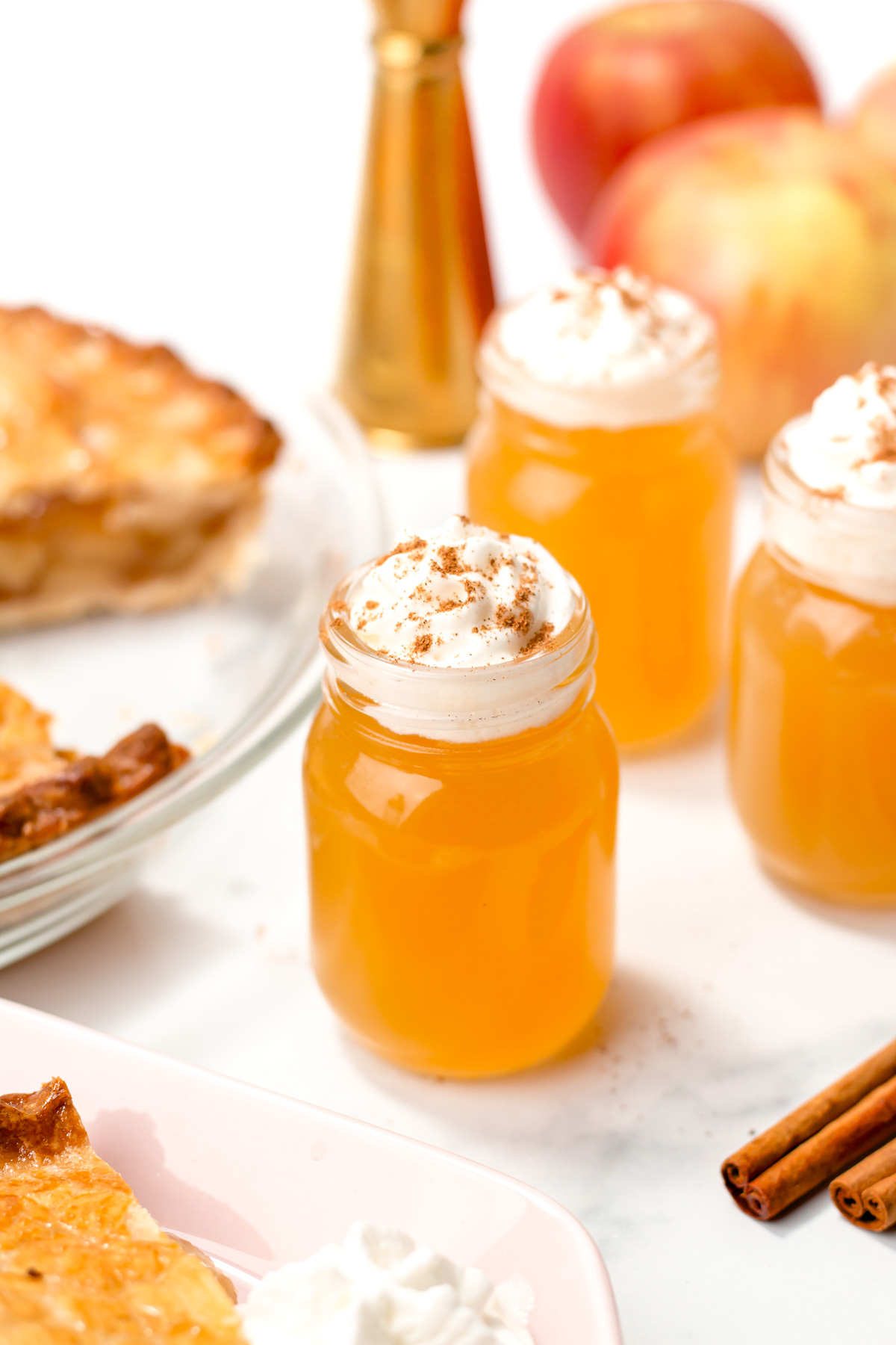 Three small mason jars filled with orange liquid, topped with whipped cream and cinnamon, sit on a white surface with apples, cinnamon sticks, and pie nearby.