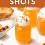 Small glass jars filled with orange apple pie shots, topped with whipped cream and cinnamon, are displayed on a white surface next to slices of pie and cinnamon sticks.