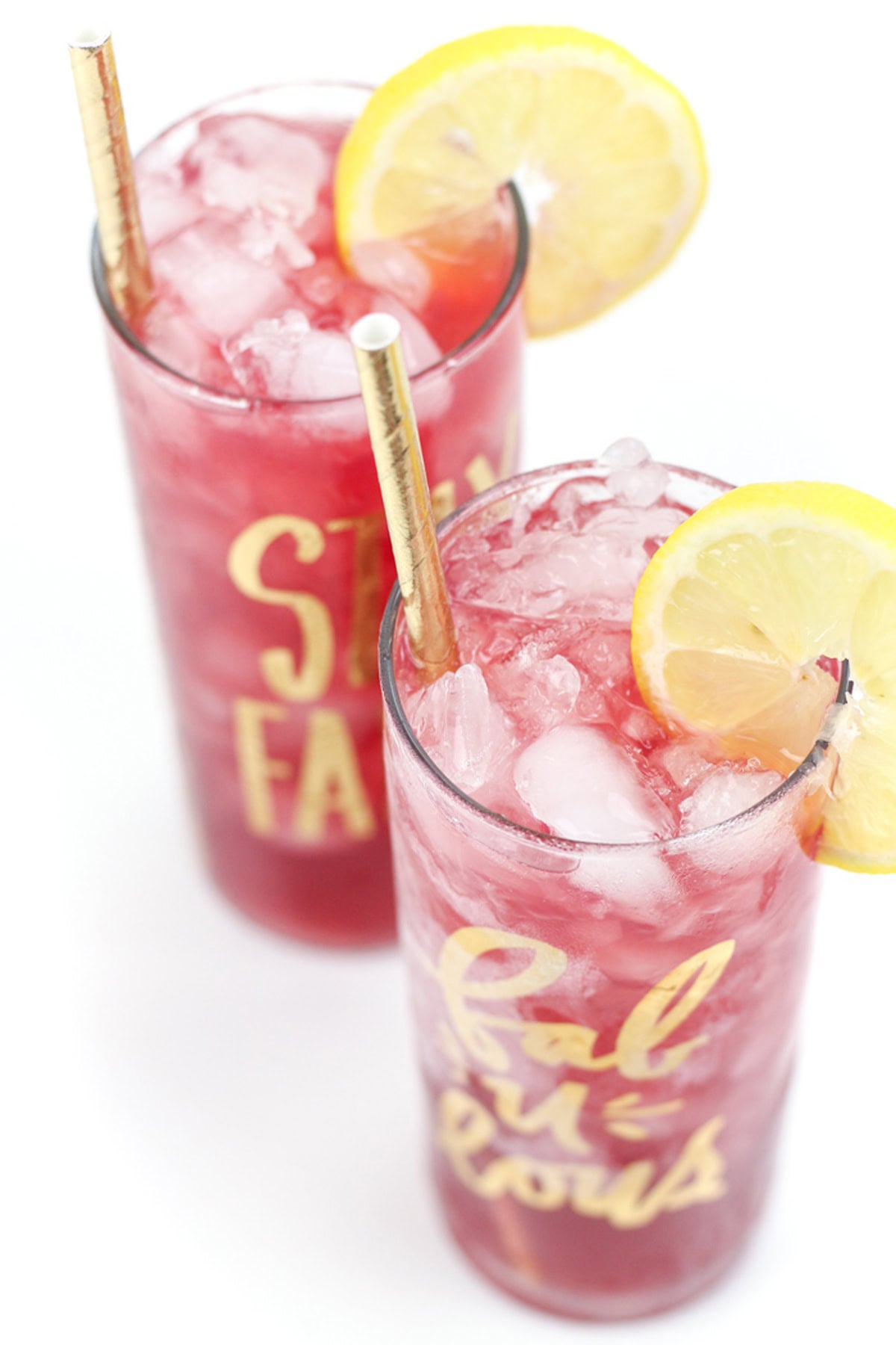 Two iced pink bourbon cocktails in tall glasses with lemon slices and gold straws, seen from above on a white background.