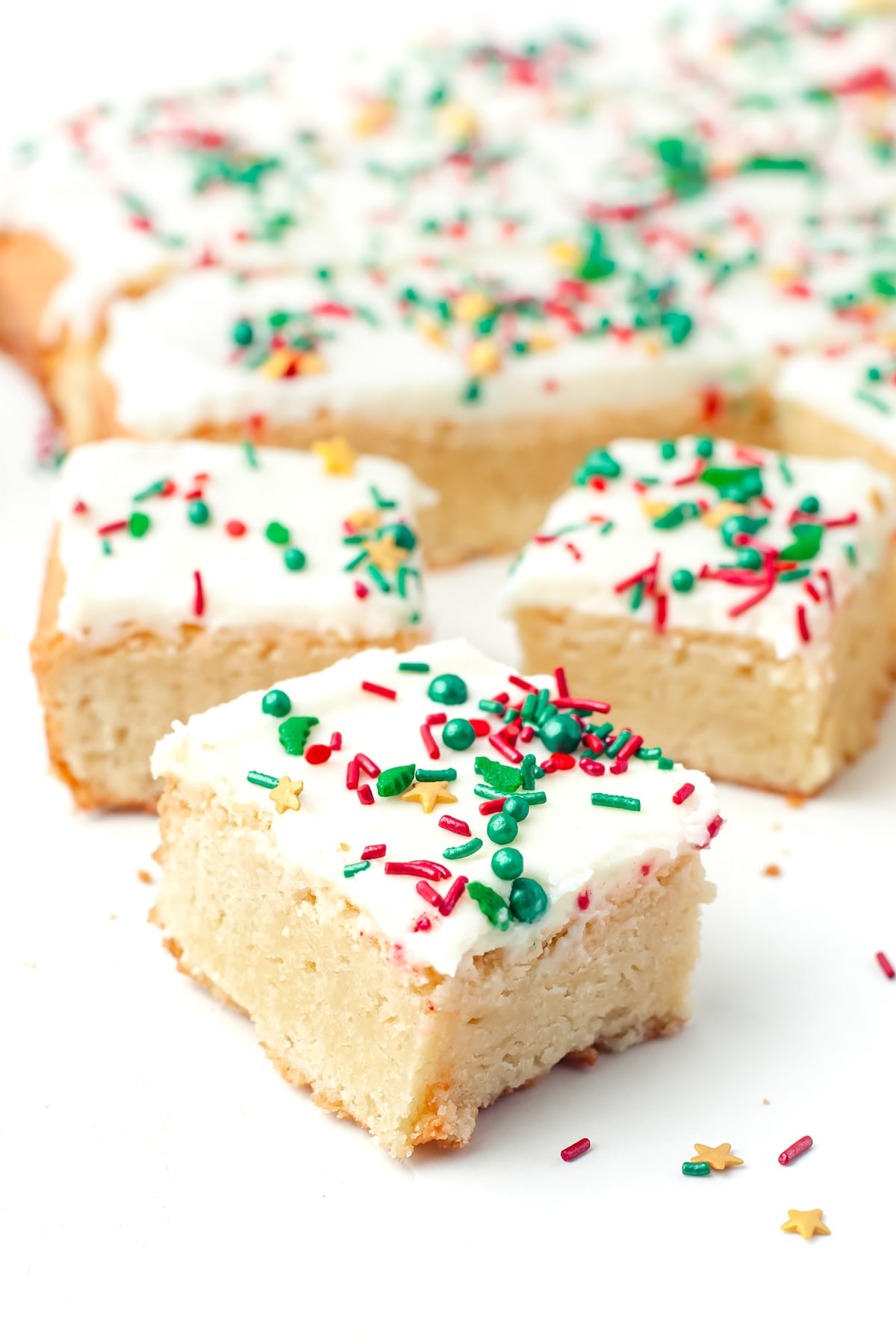Frosted sugar cookie bars topped with red, green, and yellow sprinkles are arranged on a white surface.