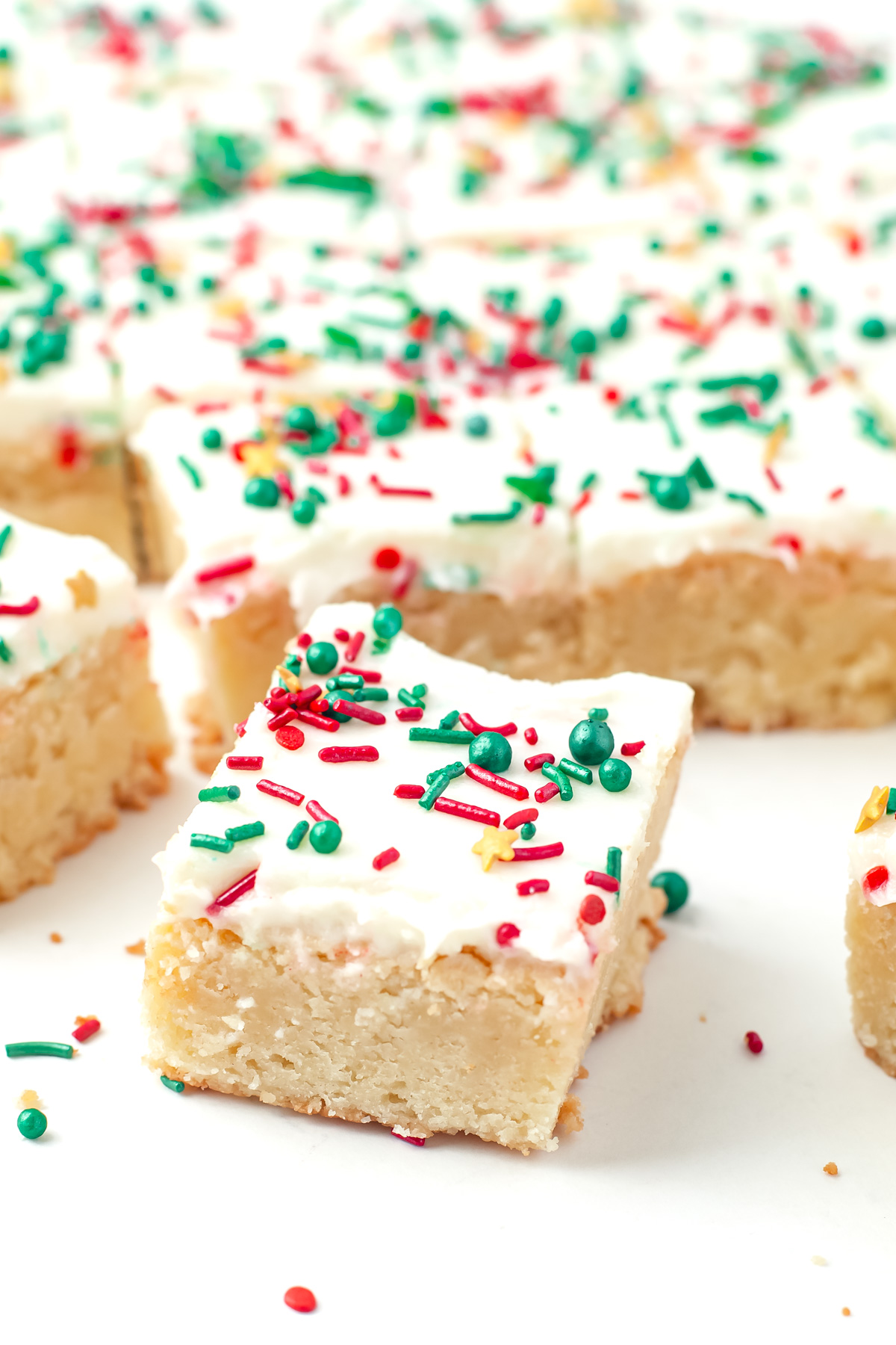 Squares of frosted blondie bars topped with white icing and red, green, and yellow holiday sprinkles on a white surface.