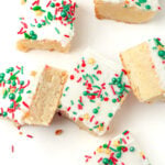 Six frosted sugar cookie bars with colorful red, green, and yellow sprinkles are arranged on a white surface, with one bar partially eaten.