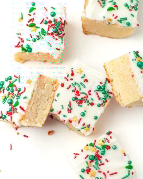 Six frosted sugar cookie bars with colorful red, green, and yellow sprinkles are arranged on a white surface, with one bar partially eaten.