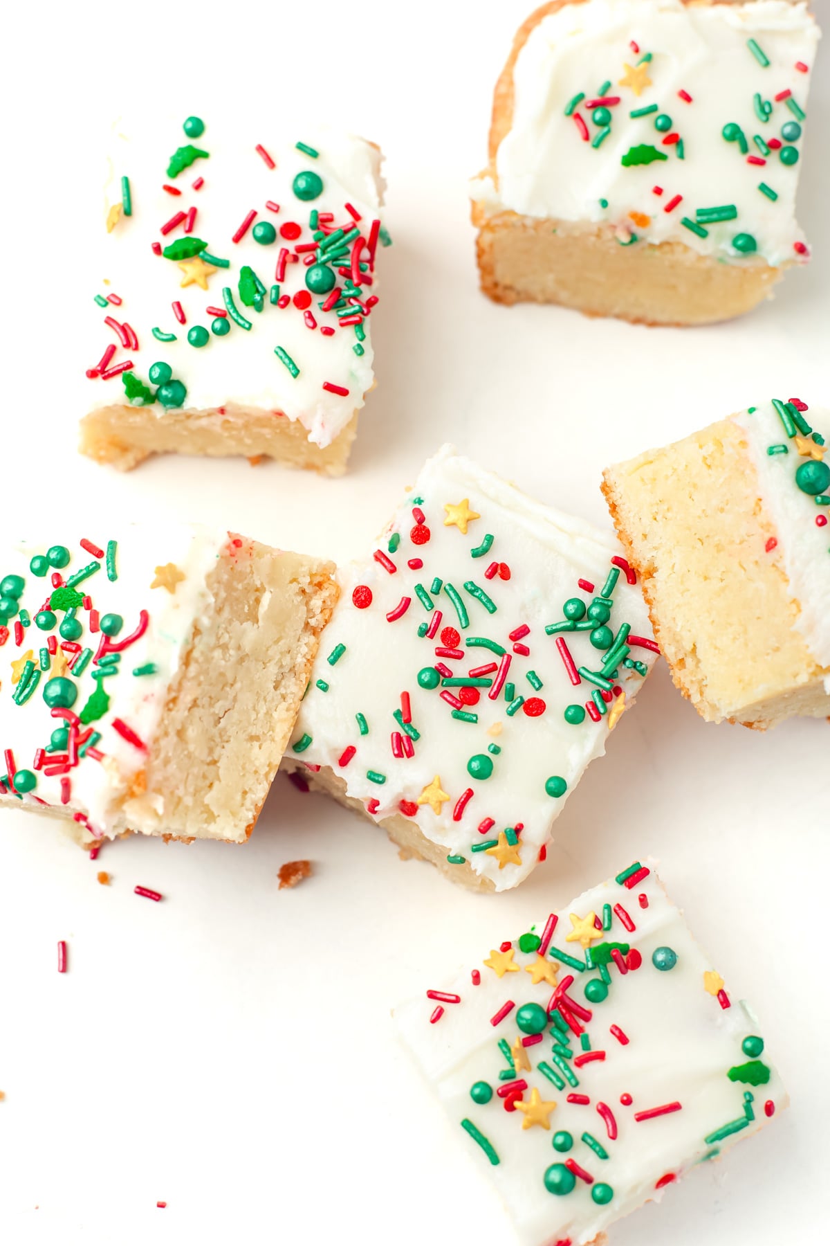 Six frosted sugar cookie bars with colorful red, green, and yellow sprinkles are arranged on a white surface, with one bar partially eaten.