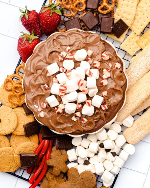 A platter with chocolate dip topped with marshmallows and peppermint pieces, surrounded by pretzels, crackers, cookies, strawberries, marshmallows, and chocolate squares.