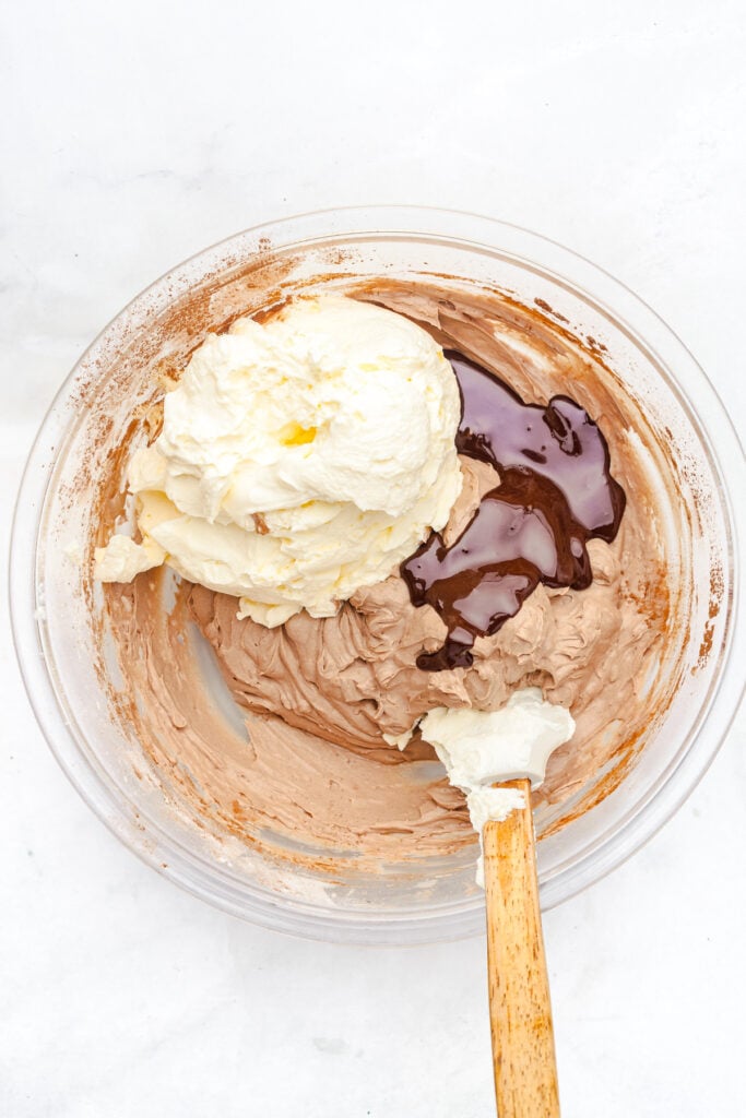 A glass bowl containing whipped cream, chocolate mixture, and partially mixed ingredients with a spatula on a white surface.