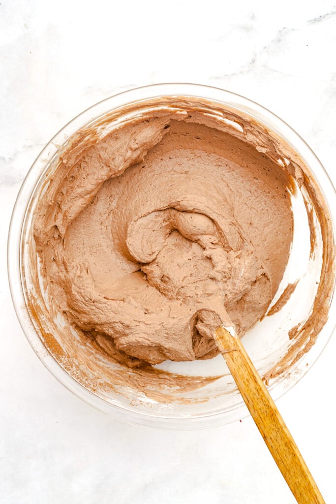 A glass bowl filled with light brown cake batter, being mixed with a wooden spoon, sits on a white surface.
