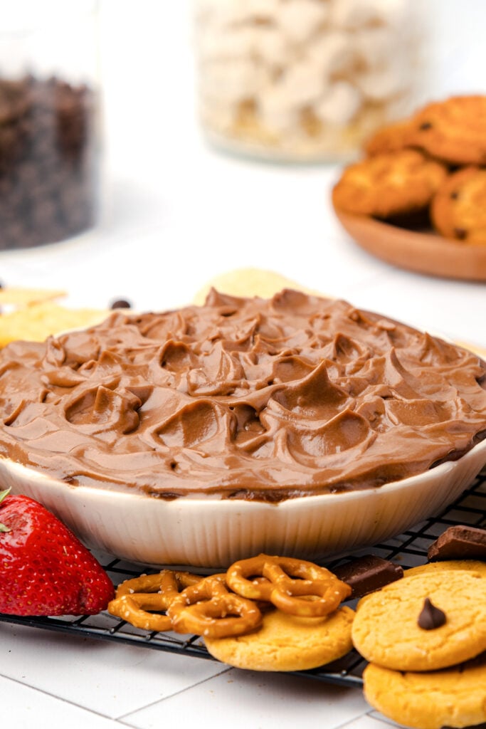 A bowl of chocolate dip surrounded by strawberries, pretzels, cookies, and chips on a cooling rack, with cookies and jars in the background.