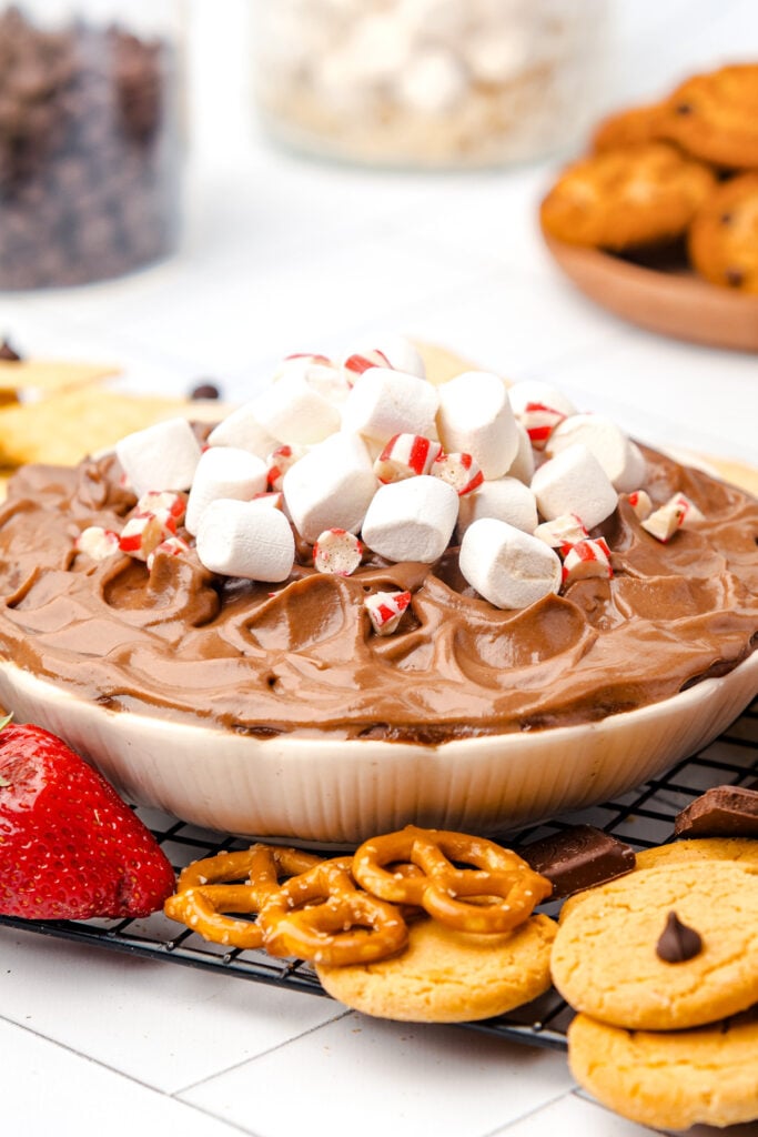 A bowl of chocolate dip topped with mini marshmallows and crushed peppermint, surrounded by strawberries, pretzels, and cookies on a wire rack.