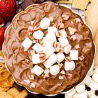 A bowl of chocolate dip topped with marshmallows and crushed peppermint, surrounded by pretzels, chocolate, cookies, graham crackers, and strawberries.