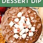 A bowl of creamy hot cocoa dip topped with mini marshmallows and crushed peppermint, surrounded by snacks for dipping.