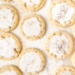 Iced sugar cookies arranged on parchment paper, topped with white glaze and sprinkled with black specks.