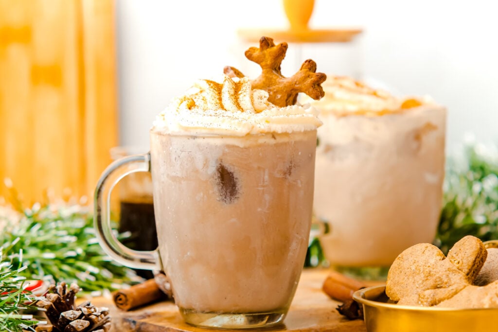 A glass mug of iced coffee topped with whipped cream and two gingerbread cookies, surrounded by holiday decorations and cookies.