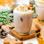 A glass mug filled with iced chocolate topped with whipped cream and a gingerbread cookie, surrounded by gingerbread cookies and festive decor.