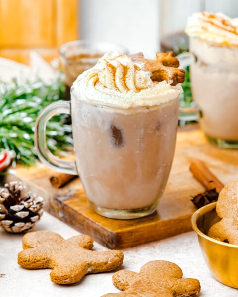 A glass mug filled with iced chocolate topped with whipped cream and a gingerbread cookie, surrounded by gingerbread cookies and festive decor.