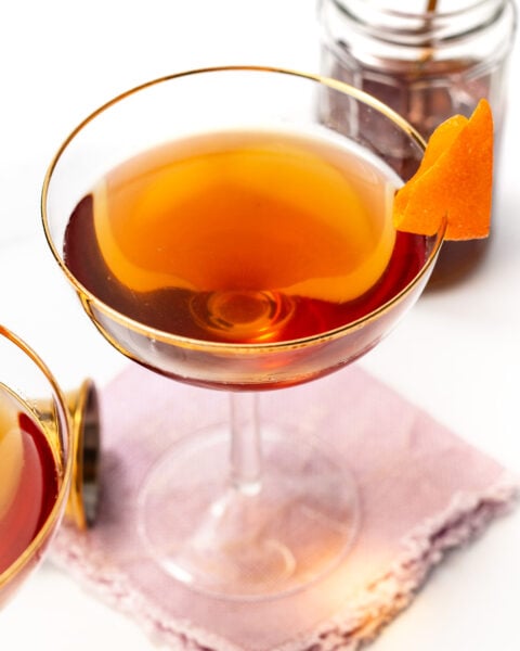 A coupe glass filled with an amber cocktail, garnished with an orange peel, sits on a purple napkin with a jar and bar spoon in the background.
