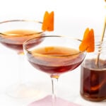 Two coupe glasses with dark brown cocktails, each garnished with an orange peel, sit on a pink napkin next to a small jar of cocktail cherries.