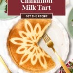 A cinnamon milk tart with a decorative flower pattern on top, a slice cut out, and gold utensils on a white plate, surrounded by flowers.