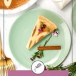 A slice of cinnamon milk tart on a green plate, garnished with a cinnamon stick and flowers, next to gold cutlery and a glass of milk.